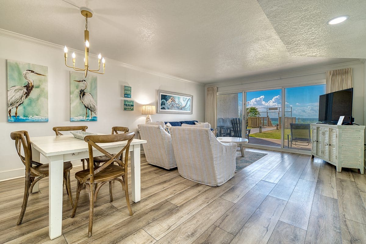 Open-concept living and dining area at The Beach House Condominiums with a wood dining table, striped chairs, and patio doors.