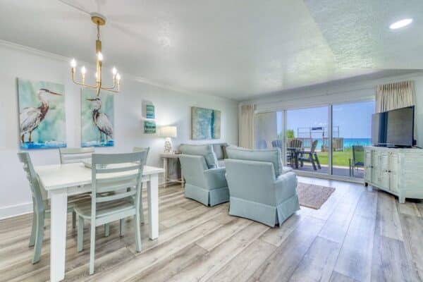 A modern living room and dining area with light blue furniture, wood flooring, large glass doors opening to a patio with an ocean view.