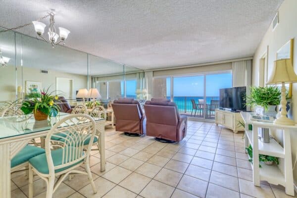 A brightly lit living room with tile flooring, two leather recliners facing a TV, a dining table with chairs, and large windows overlooking a balcony with ocean views.