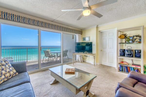 Beachfront condo lounge with wide windows, ocean view, furnished with a sofa, chair, TV and bookshelf at The Beach House.
