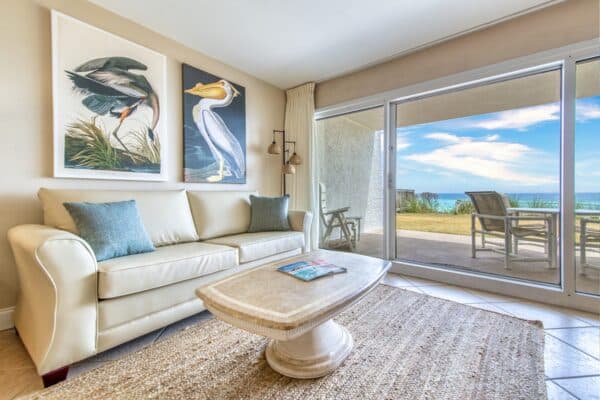 Bright living room with white sofa, glass table, bird paintings and open door to patio at Beach House Condominiums.