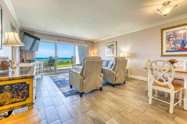 A living room with two armchairs, wall-mounted TV, and dining area. A sliding glass door leads to a balcony with ocean views.