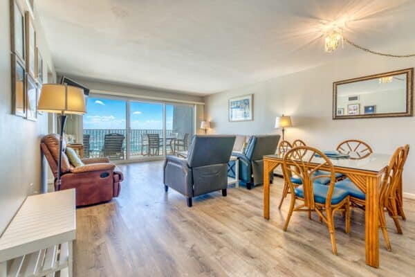 Living area with a tan recliner, two dark armchairs, wooden dining table with chairs, wall-mounted TV, balcony with ocean view. Wood flooring and ceiling lights.