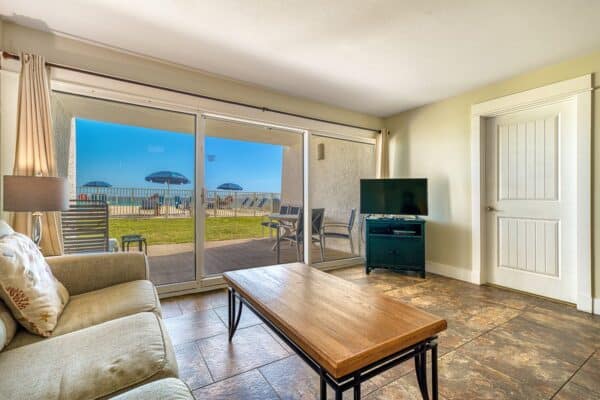 Living room with sofa, coffee table, TV, tile floor, and glass doors to a patio overlooking the ocean and beach umbrellas.