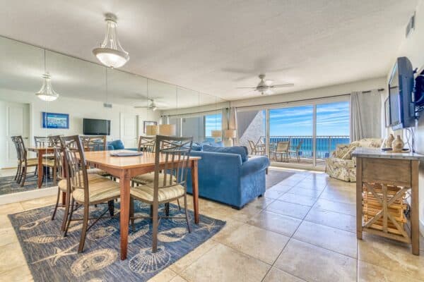 Open living area with dining table, blue sofa, armchair, wall-mounted TV, mirrors, and glass doors to balcony. Ocean view included.