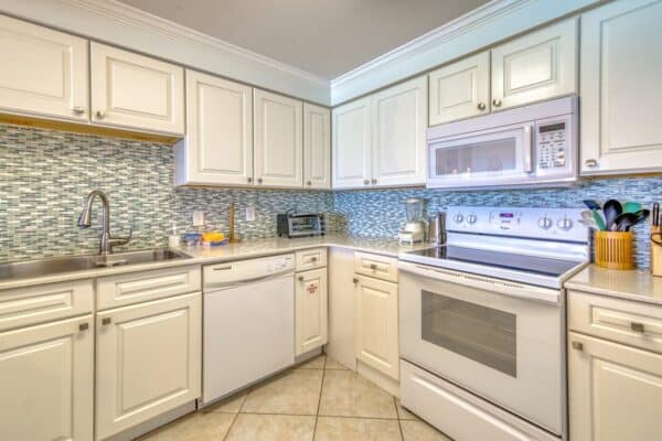 A modern kitchen with white cabinets, tiled backsplash, dishwasher, microwave, oven, and countertop with cooking utensils.