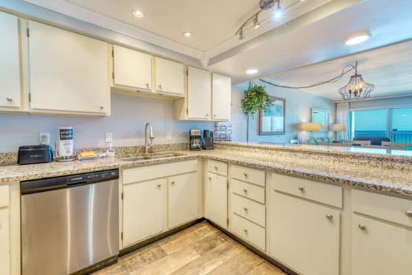 A modern kitchen with beige cabinets, granite countertops, stainless steel dishwasher and sink. Toaster and coffee maker on the counter.