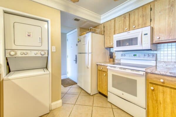 Compact kitchen at The Beach House Condominiums with wooden cabinets and white appliances, adjacent to a narrow corridor.