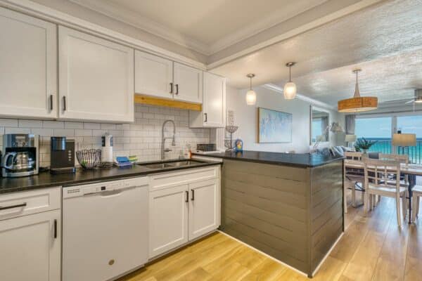 Modern kitchen with white cabinets, black countertops, subway tiles, and ocean view through large dining area windows.
