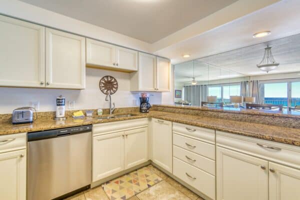 Kitchen with white cabinets, granite countertops, stainless steel dishwasher, and toaster; view of living room and sea.