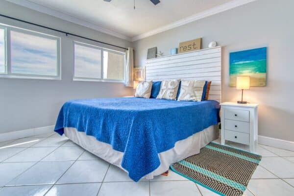 Bright bedroom at The Beach House Condominiums with large bed, blue cover, white 'sleep' headboard, nightstand, striped rug and coastal wall art.