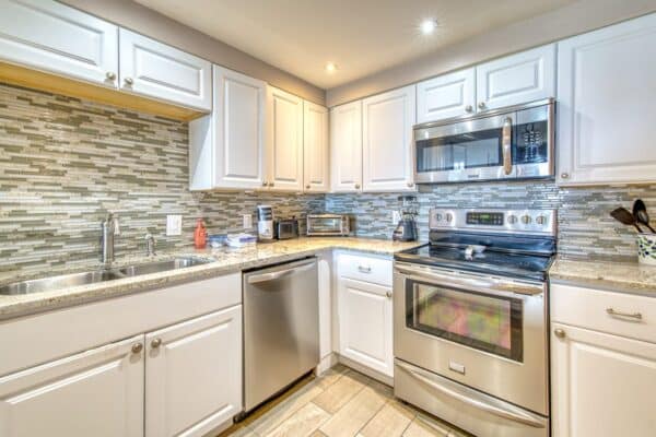 Modern kitchen with stainless steel appliances, white cabinets, mosaic tile backsplash, granite countertops, and light wood flooring.