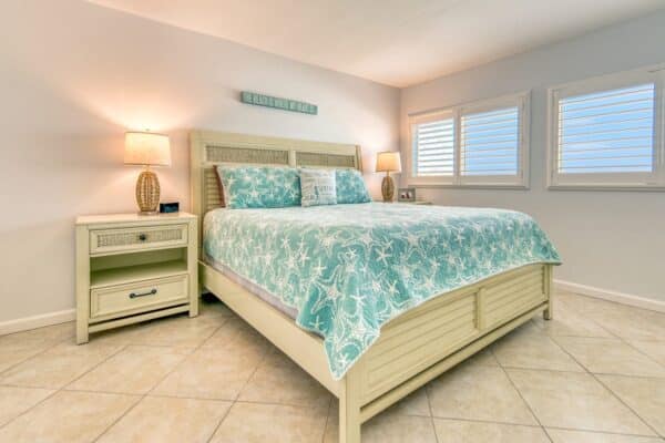 A bedroom with a double bed, teal seashell-patterned bedding, bedside tables with lamps, and a shuttered window. A wall sign says "Relax. You're At The Beach.