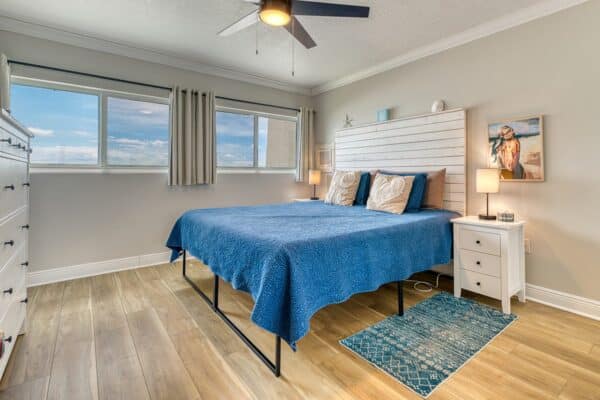Bedroom with blue bedspread, two nightstands, lamps, ceiling fan, large windows, wood floor, and coastal decor.