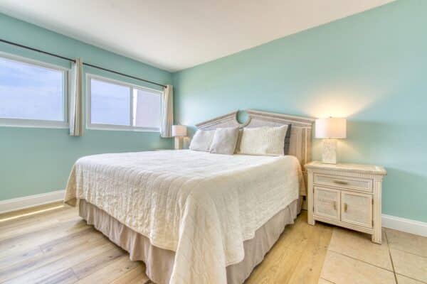 Bright bedroom in Beach House Condominiums with a large bed, white bedspread, teal walls, windows and hardwood floor.