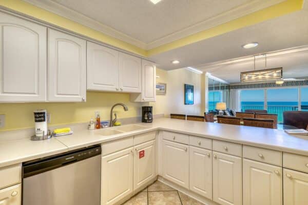 Bright kitchen with white cabinets, stainless dishwasher, coffee maker, and open view to living area with ocean-facing windows.