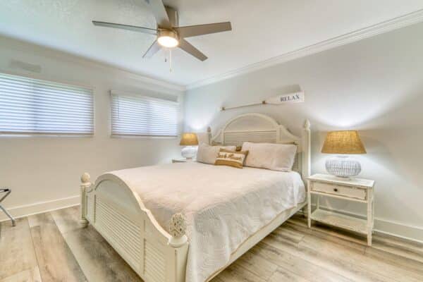 A B105 bedroom with a large white bed, two nightstands with lamps, window blinds, a ceiling fan, and wooden floors. "RELAX" sign and "Beach House" pillow visible.