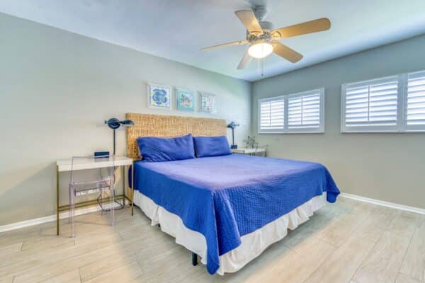 A clean bedroom with a blue bedspread, ceiling fan, rattan headboard, two nightstands, framed pictures above the bed, and windows with white plantation shutters.