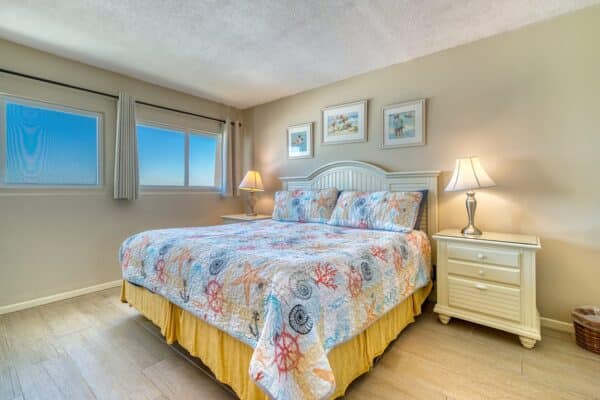 A colorful seashell comforter covers a neatly made bed with lamps, nightstands, art above the headboard, and curtained window.