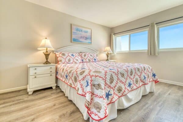 A bedroom with a large bed, seashell quilt, bedside tables with lamps, and a window with light curtains. Room B402.