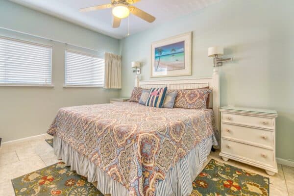 A neatly made bed with patterned bedding, flanked by white nightstands and lamps. A beach-themed picture hangs above.