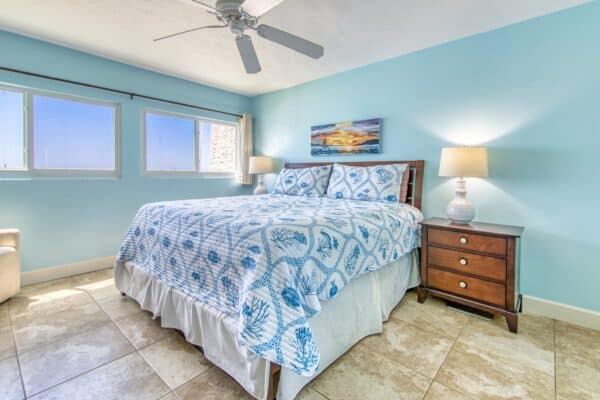 Bright bedroom at The Beach House Condos with queen bed, blue-white bedding, wooden nightstand, lamp, ceiling fan, bell, and painting.