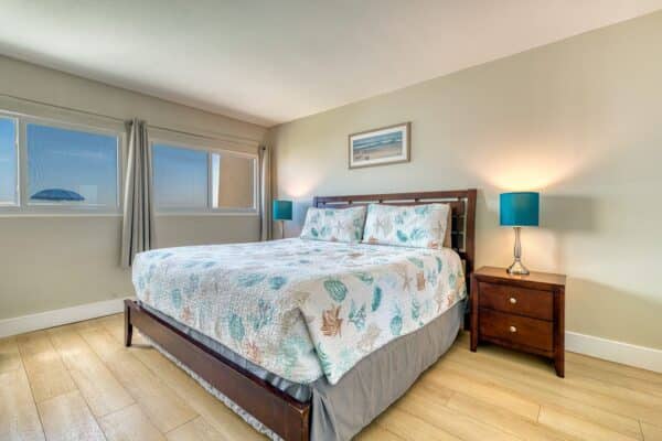Bedroom with large bed, two bedside tables with turquoise lamps, light wood floor, and two curtained windows.