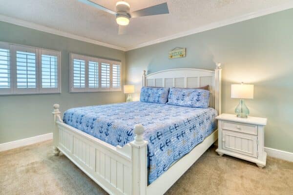 Bedroom with white wooden bed, blue anchor bedding, two nightstands, ceiling fan, carpeted floor, and three shuttered windows.
