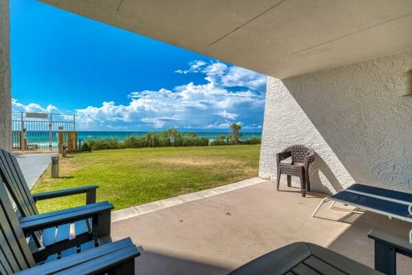 Covered patio with chairs and a lounge chair, overlooking a grassy lawn and the ocean at the condominium complex.