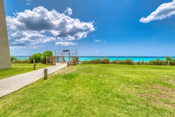 A grassy area with a wooden path leading to a covered gate marked B105, with the ocean and a partly cloudy sky in the background.