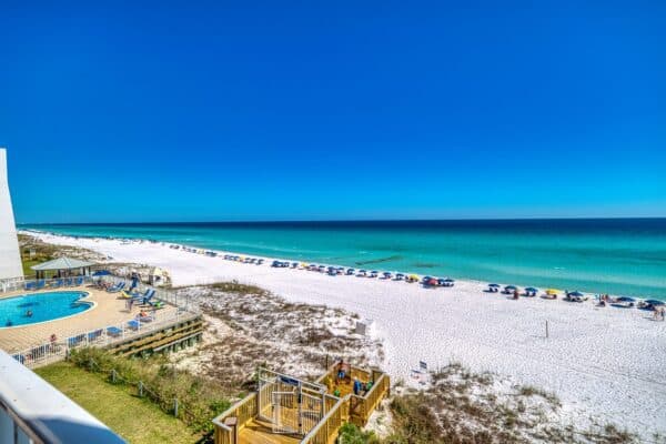 Beachfront view of white sand, turquoise water, beach umbrellas, and a pool near the shore under a clear blue sky.