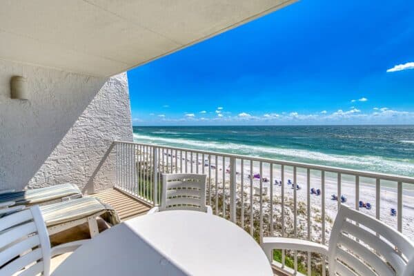 B402: Outdoor balcony with white furniture overlooking a sandy beach and calm ocean waves under a clear blue sky.