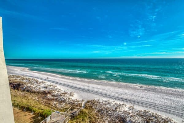 View of a sandy beach with a clear blue ocean and sky. A path runs parallel, with a small wooden deck on the left.