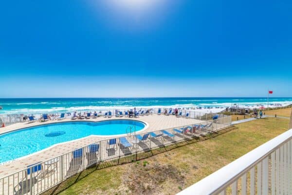 Balcony view of a pool and crowded beach at The Beach House Condominiums on a sunny day with clear skies and blue ocean waves.