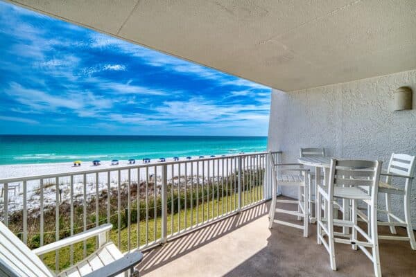 Balcony with white chairs and table overlooks ocean, sandy beach, and colorful umbrellas under a blue sky with clouds.