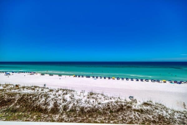 A wide sandy beach near the condos with colorful umbrellas, turquoise water, and people swimming under a blue sky.