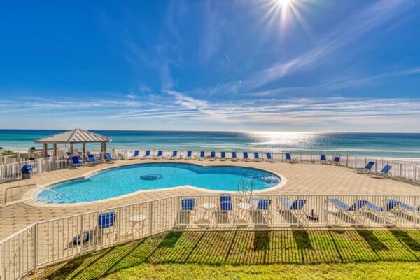 A circular outdoor pool with blue lounge chairs and a white fence, overlooking a beach and ocean.