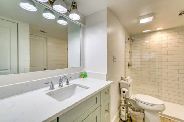 A modern bathroom with a white vanity, under-mount sink, large mirror, overhead lights, and a glass-door shower next to the toilet.