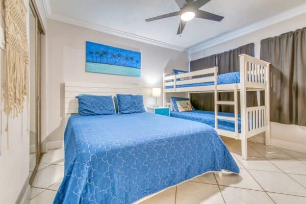 A203 at Beach House Condos featuring a bright bedroom with a large blue bed, white bunk bed, and beach-themed painting.