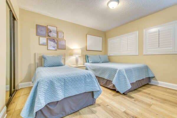 Bright bedroom at The Beach House Condominiums with two single beds in blue covers, wall art, and wooden floors.