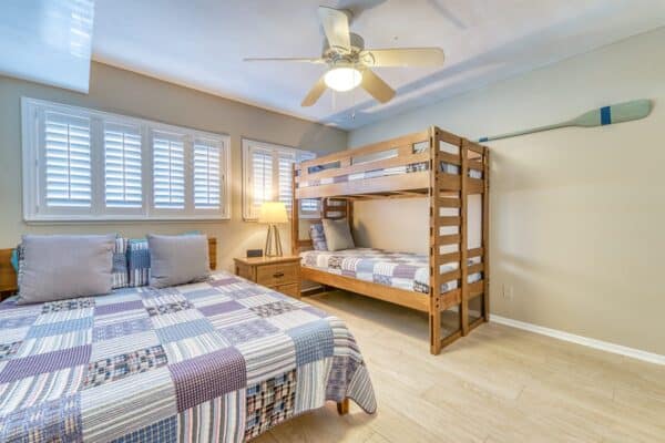 A bedroom, labeled C302, with a ceiling fan, queen bed, wooden bunk bed, beige walls, nightstand with lamp, and blue and white oar on the wall.