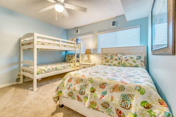 A bedroom with a ceiling fan, light blue walls, beige carpet, a bunk bed, and a queen bed with sea-themed bedding. Two windows with blinds.