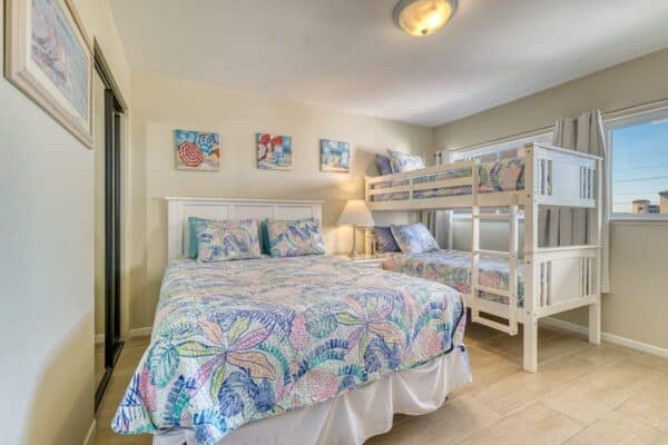 Bright bedroom at the condominium with queen and bunk beds, colorful bedding, wood floors, wall art, and natural light.