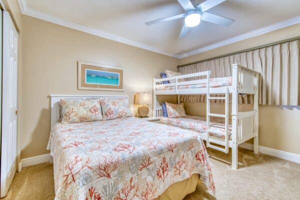 Well-lit room at the Beach House Condominiums with a double bed, bunk bed, steel ceiling fan and framed artwork.