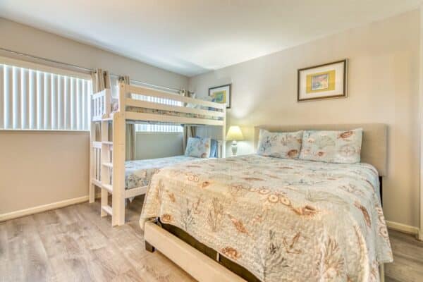 A bedroom with a large bed, bunk bed, two framed pictures, a lamp on a bedside table, and a window with vertical blinds.