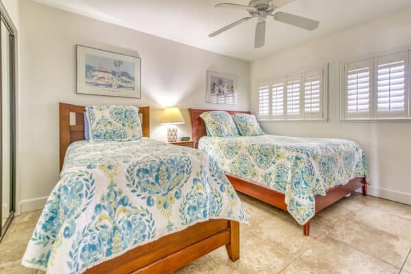 Bedroom with two wooden beds and floral bedding, lamp on a nightstand, shuttered window, and ceiling fan overhead.