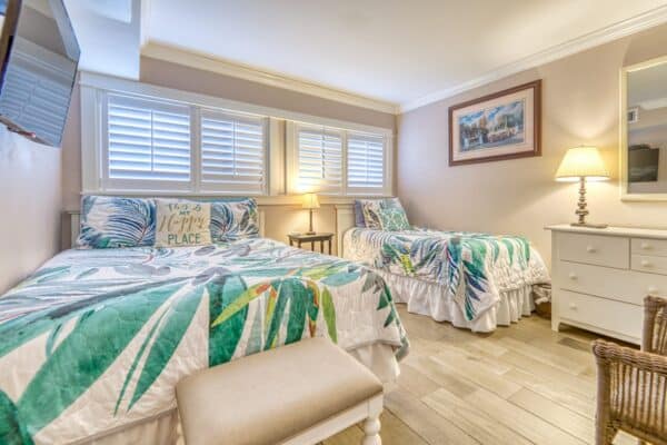 A bedroom labeled B101 with two beds featuring tropical-themed bedding, a painting on the wall, a dresser with a lamp, and shutters allowing natural light.