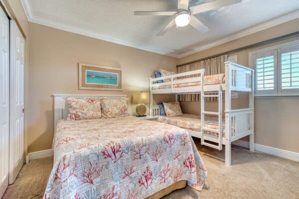 Bedroom at the condo with double bed, white bunk beds, coral bedding, ceiling fan, lamp, framed art, and shuttered window.