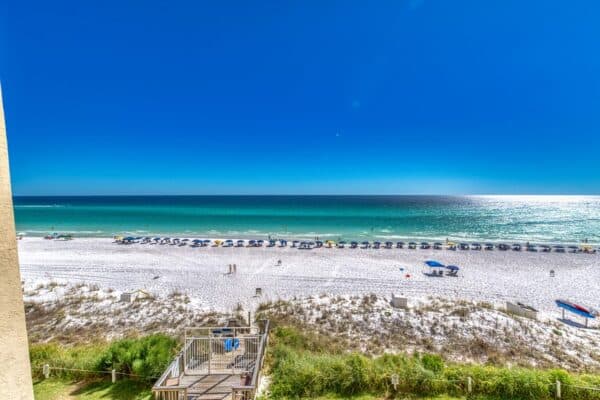 A clear beach day with calm blue water, white sand, and rows of blue beach umbrellas marked B405. People walk and relax by a wooden deck.