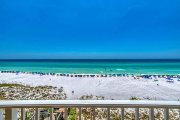 A sandy beach with blue umbrellas near the shoreline, people swimming in the turquoise ocean under a clear blue sky, seen from behind a white railing.